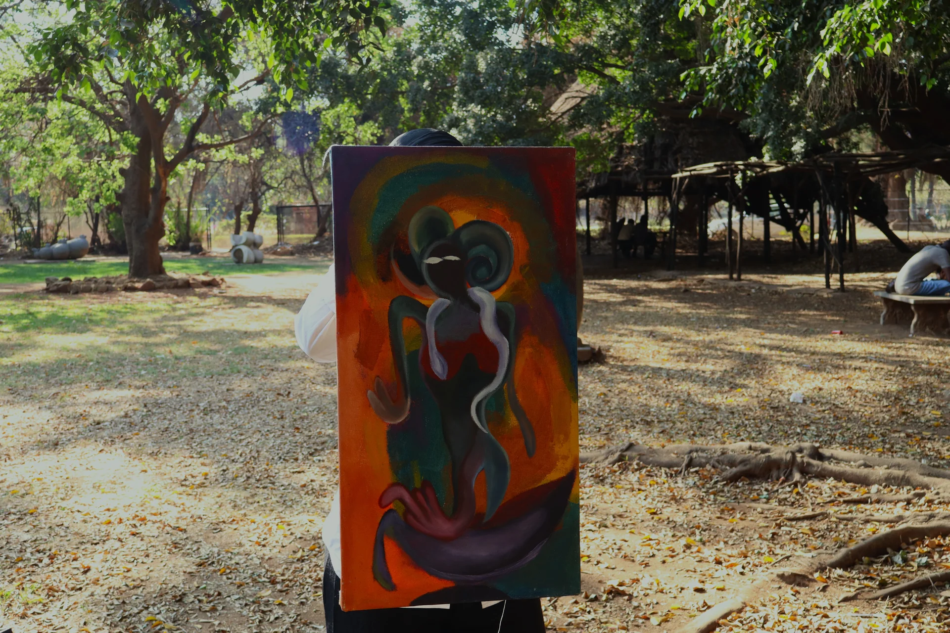 A person stands outdoors in a garden holding a large abstract painting featuring a dark spiritual figure against bright orange and green tones.