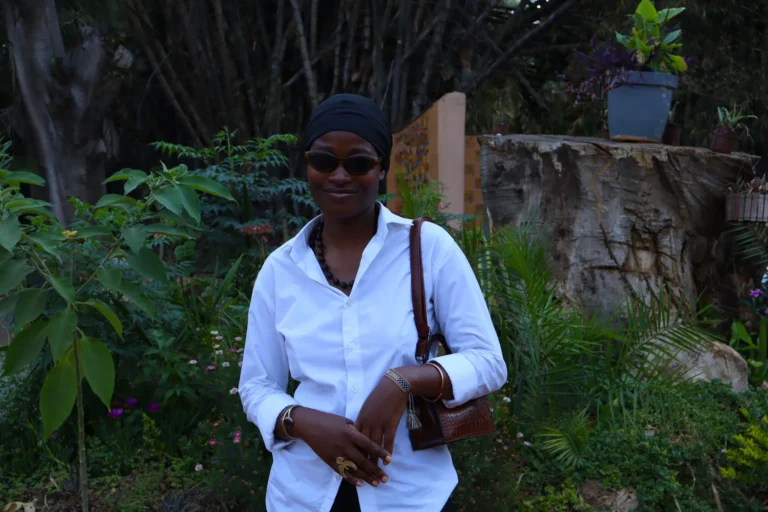 A young woman wearing a white shirt, headwrap, and sunglasses stands in a lush garden setting, smiling softly.